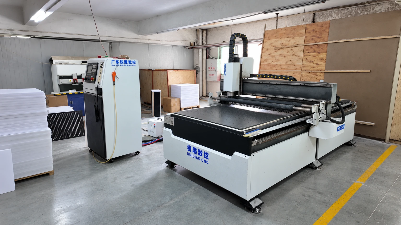 Flatbed CNC router with a separate control cabinet and stacked sheets in an acrylic fabrication workshop